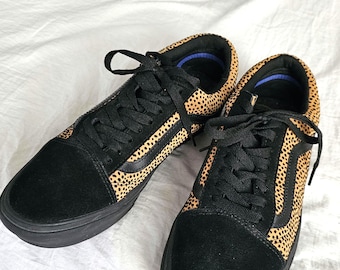 May include: A pair of black and tan sneakers with a cheetah print design. The sneakers have black laces, a black sole, and a black suede toe and heel. The interior is blue.