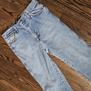 May include: Light blue denim jeans with a classic five-pocket design. The jeans have a button and zipper closure, belt loops, and a faded wash. The garment is laid flat on a wooden surface.