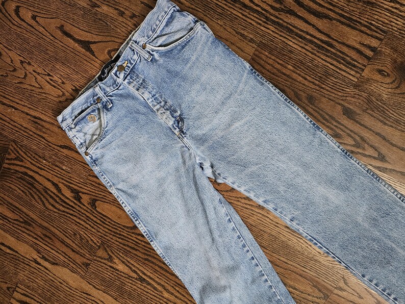 May include: Light blue denim jeans with a classic five-pocket design. The jeans have a button and zipper closure, belt loops, and a faded wash. The garment is laid flat on a wooden surface.