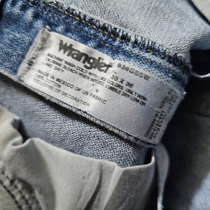 May include: Close-up of a light blue Wrangler denim jeans label. The label displays the brand name, size 33 x 36, and care instructions. The jeans are made in Mexico of US fabric and are 100% cotton.