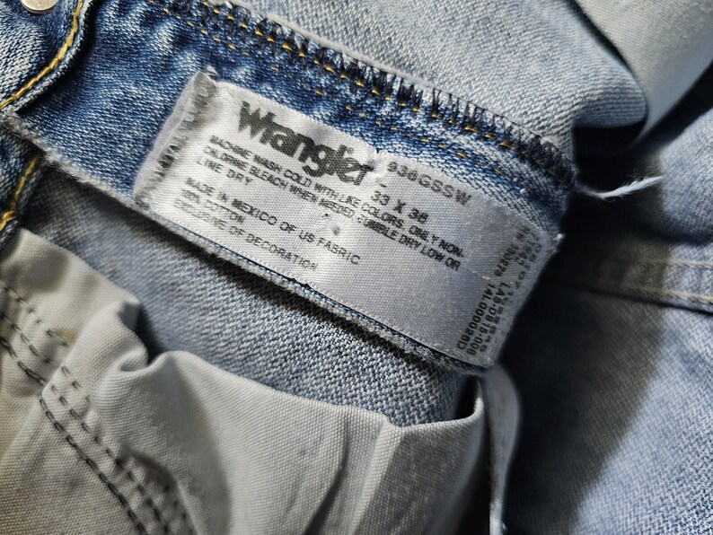 May include: Close-up of a light blue Wrangler denim jeans label. The label displays the brand name, size 33 x 36, and care instructions. The jeans are made in Mexico of US fabric and are 100% cotton.