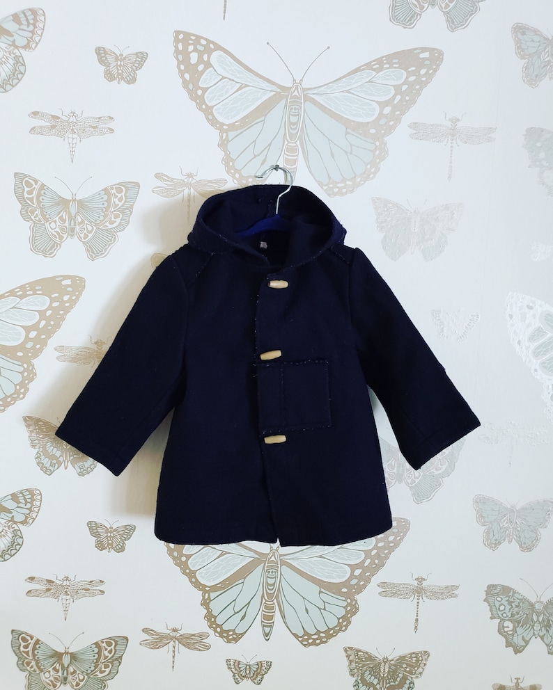 May include: A navy blue hooded coat with wooden toggle closures and a patch pocket on the front. The coat is hanging on a hanger against a white background with a butterfly pattern.