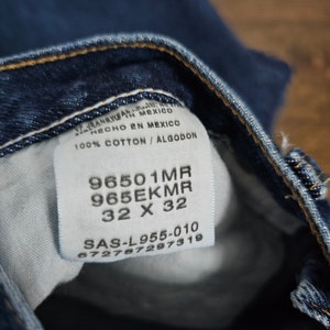 May include: A close-up of a blue denim jean label with white text. The label includes the text "96501MR", "965EKMR", "32 X 32", "SAS-L955-010", and "672787297319".