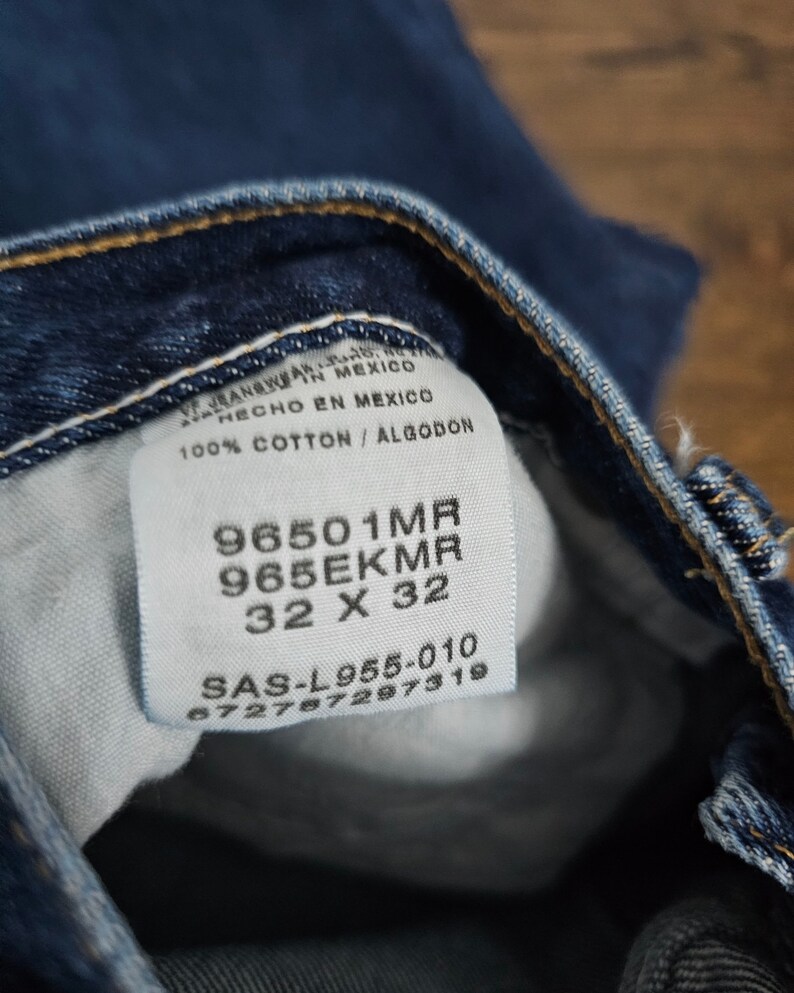 May include: A close-up of a blue denim jean label with white text. The label includes the text "96501MR", "965EKMR", "32 X 32", "SAS-L955-010", and "672787297319".