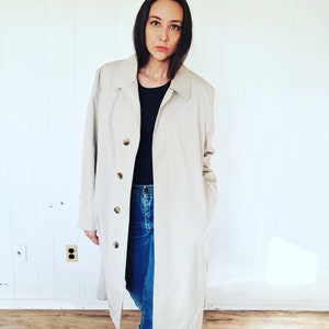 May include: A beige trench coat with a button-up front and a collared neckline. The coat is worn over a black top and blue jeans. The background is a white wall.
