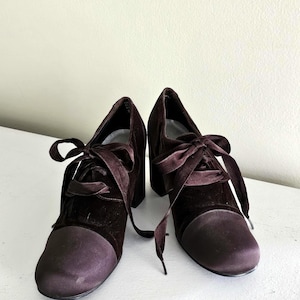 May include: A pair of dark brown velvet heeled shoes with satin toe caps and matching satin laces. The shoes have a chunky heel and a lace-up closure.