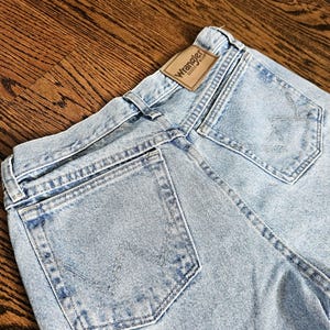 May include: Light blue denim jeans with a classic five-pocket design. The Wrangler brand label is visible on the back waistband. The jeans have a faded wash and are made of durable denim material.