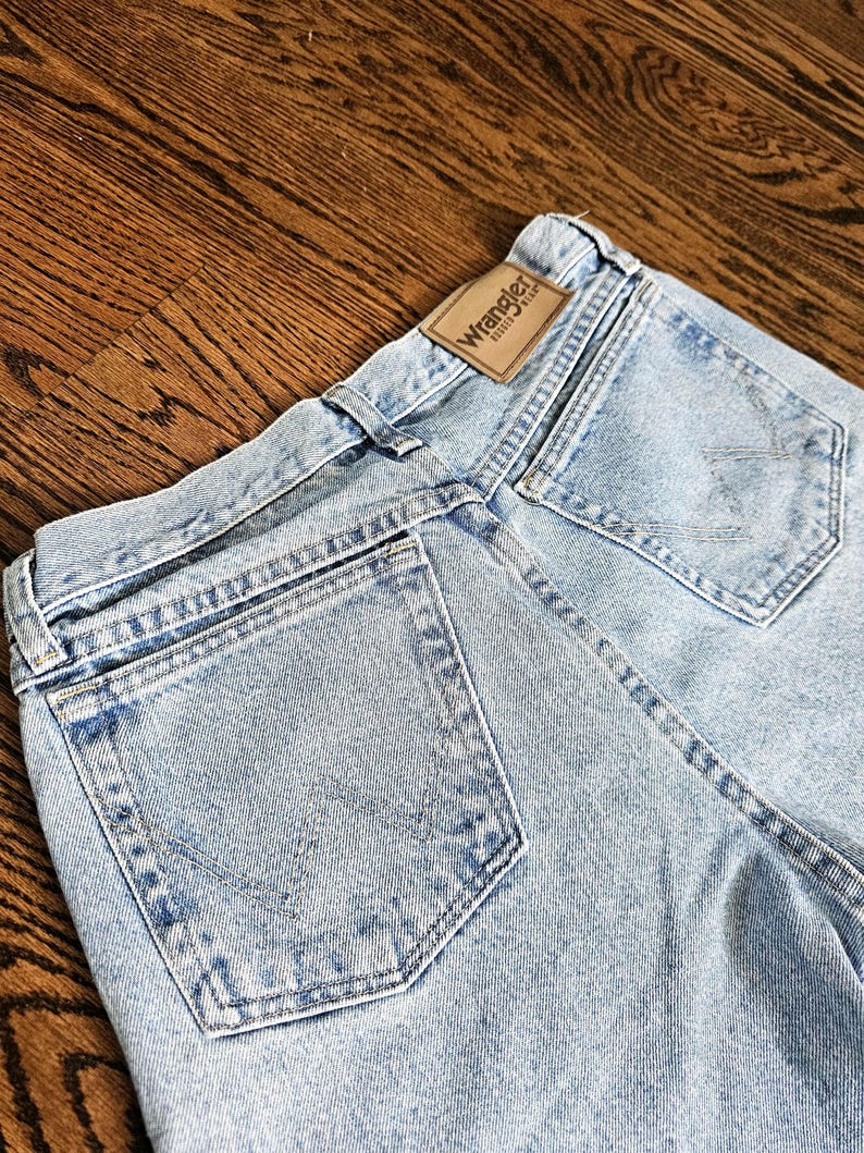 May include: Light blue denim jeans with a classic five-pocket design. The Wrangler brand label is visible on the back waistband. The jeans have a faded wash and are made of durable denim material.