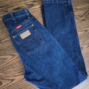 以下が含まれることがあります： 後ろのポケットに「Wrangler」と書かれた茶色の革のパッチが付いた濃い青色のデニムジーンズです。
