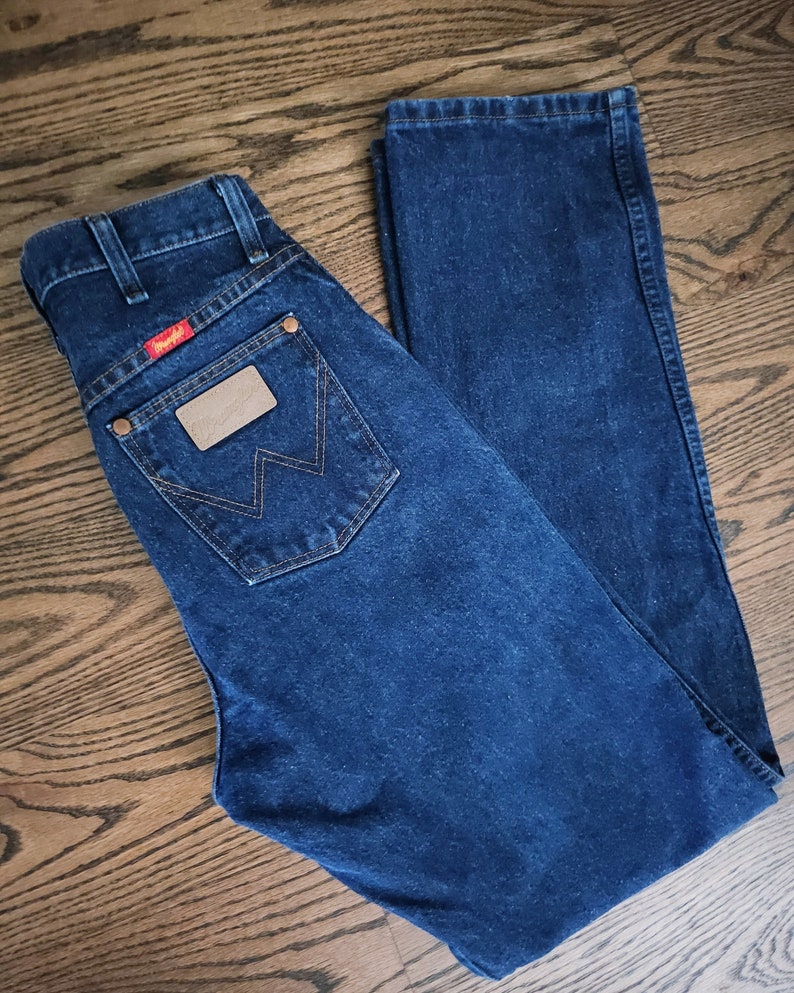 以下が含まれることがあります： 後ろのポケットに「Wrangler」と書かれた茶色の革のパッチが付いた濃い青色のデニムジーンズです。