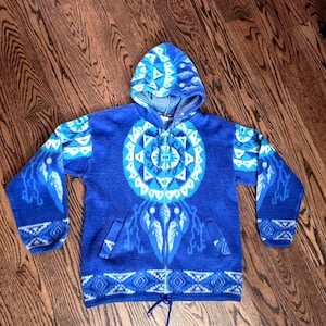 May include: A bright blue fleece zip-up hoodie featuring a large circular geometric pattern on the chest and sleeves. The garment includes a drawstring hood, two front pockets, and a drawstring hem with blue cords, showcasing a repeating diamond motif.