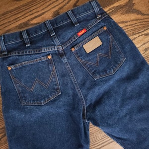 以下が含まれることがあります： 後ろのポケットに「Wrangler」と書かれた茶色の革のパッチが付いた濃い青色のデニムジーンズです。