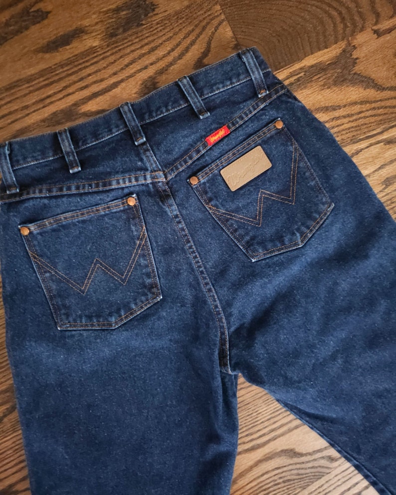 以下が含まれることがあります： 後ろのポケットに「Wrangler」と書かれた茶色の革のパッチが付いた濃い青色のデニムジーンズです。
