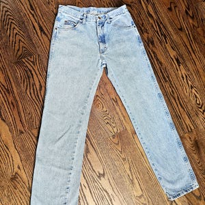 May include: Light blue denim jeans with a classic five-pocket design. The jeans have a straight leg cut and a button and zipper closure. The denim has a faded wash, giving them a vintage look. The jeans are laid flat on a wooden surface.
