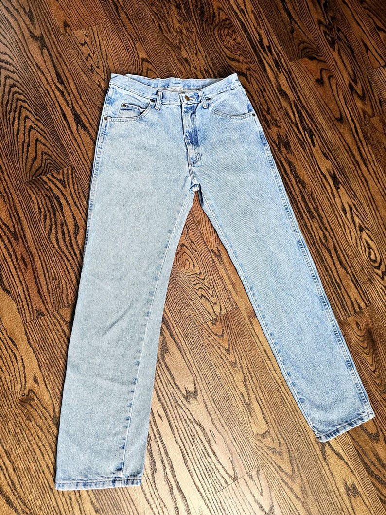 May include: Light blue denim jeans with a classic five-pocket design. The jeans have a straight leg cut and a button and zipper closure. The denim has a faded wash, giving them a vintage look. The jeans are laid flat on a wooden surface.