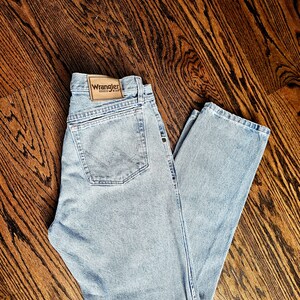 May include: Light blue denim jeans with a classic five-pocket design. The jeans feature a Wrangler brand patch on the back and are laid flat on a wooden surface. The jeans have a straight leg cut.
