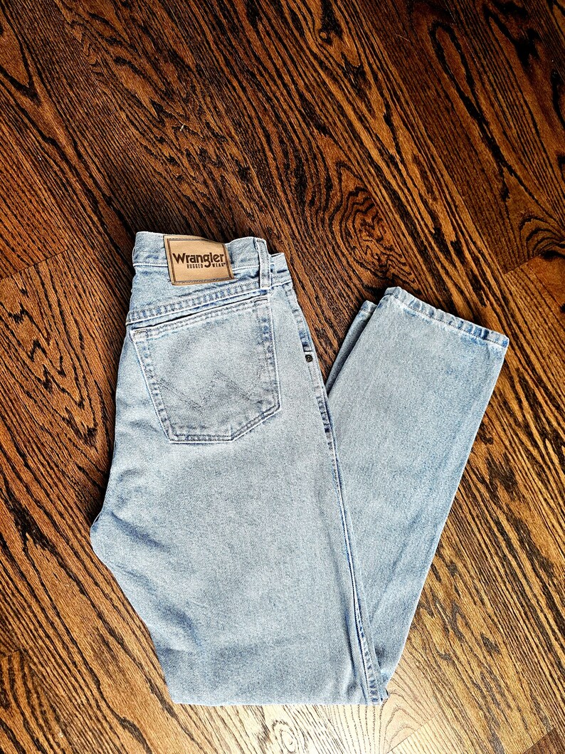 May include: Light blue denim jeans with a classic five-pocket design. The jeans feature a Wrangler brand patch on the back and are laid flat on a wooden surface. The jeans have a straight leg cut.