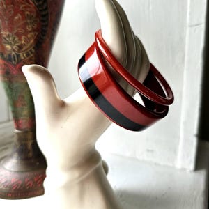 May include: Two red and black striped bangles are displayed on a cream-colored hand-shaped display. The bangles have a glossy finish. A red and gold decorative vase is in the background. The bracelets are a vintage accessory.