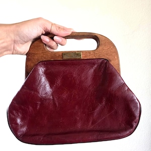 May include: A deep burgundy leather handbag featuring a smooth, arched wooden handle. A rectangular brass-toned metal plaque is centered on the wooden frame, displaying engraved text. The leather body has a soft, creased texture and a wide base.