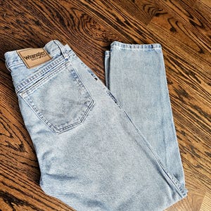 May include: Light blue Wrangler jeans with a classic five-pocket design. The jeans feature a leather patch with the Wrangler logo on the back waistband. The denim has a faded wash, giving it a vintage look.