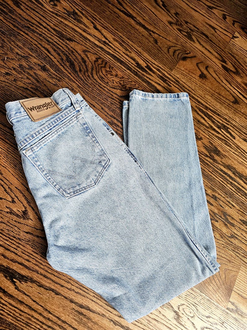 May include: Light blue Wrangler jeans with a classic five-pocket design. The jeans feature a leather patch with the Wrangler logo on the back waistband. The denim has a faded wash, giving it a vintage look.
