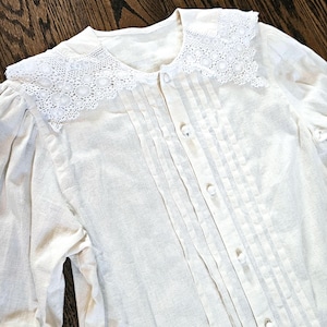 May include: Cream-colored vintage blouse with a delicate lace collar and vertical pleats down the front. The long-sleeved shirt features small, round buttons and a soft, textured fabric. The blouse is a classic piece of apparel.