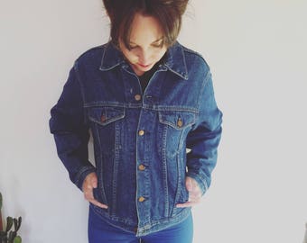 Puede incluir: Una chaqueta vaquera azul oscuro con cierre de botones y dos bolsillos delanteros. La chaqueta la lleva puesta una persona con el pelo castaño y está de pie frente a una pared blanca.