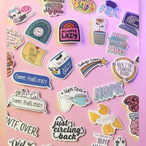 Op de afbeelding: Een verzameling kleurrijke stickers met diverse ontwerpen en tekst op een roze achtergrond. De stickers bevatten zinnen als "Always Lazy", "Damn, that's crazy" en "Sips tea". Andere ontwerpen zijn een regenboog, een laptop en een melkpak.
