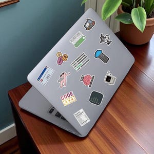 K&ouml;nnte beinhalten: Ein grauer Laptop mit verschiedenen Aufklebern, darunter "ENGINEER MODE ON", Code-bezogene Grafiken und der Text "I HATE PROGRAMMING". Der Laptop steht auf einer Holzoberfl&auml;che, im Hintergrund eine Topfpflanze.