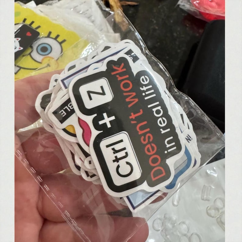 K&ouml;nnte beinhalten: Eine transparente Plastikt&uuml;te gef&uuml;llt mit verschiedenen Aufklebern. Die Aufkleber zeigen ein schwarz-wei&szlig;es Design mit dem Text "Ctrl + Z Doesn't work in real life" in Rot und Wei&szlig;. Weitere Aufkleber sind sichtbar.