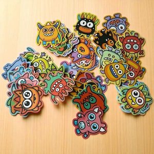 Pode incluir: Uma coleção de autocolantes de monstros coloridos com vários designs. Os autocolantes apresentam monstros de desenhos animados em tons de laranja, amarelo, azul, verde e roxo. Cada autocolante tem uma expressão e forma únicas, com uma estética lúdica e caprichosa.