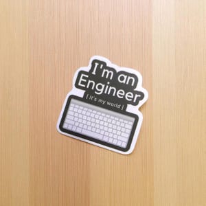K&ouml;nnte beinhalten: Ein wei&szlig;er Aufkleber mit dem Text "I'm an Engineer" in Schwarz, &uuml;ber einer Tastatur-Grafik. Der Text "[It's my world]" befindet sich unter der Tastatur. Der Aufkleber liegt auf einer hellbraunen Holzoberfl&auml;che.