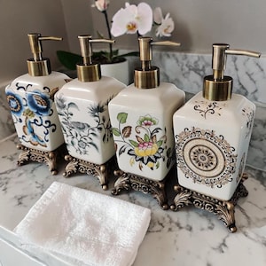 Könnte beinhalten: Vier Keramik-Seifenspender mit floralen Mustern in Blau, Grün und Gelb, jeweils mit einer bronzefarbenen Pumpe und einem dekorativen Ständer. Ein weißes Waschtuch liegt vor den Spendern.