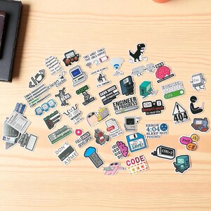 K&ouml;nnte beinhalten: Eine Sammlung bunter Aufkleber mit verschiedenen computerbezogenen Grafiken und Texten. Designs umfassen Computersymbole, Fehlermeldungen und S&auml;tze wie "Engineer in Progress" und "Dark Mode". Die Aufkleber sind auf einer hellen Holzoberfl&auml;che angeordnet.