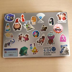 K&ouml;nnte beinhalten: Ein silberner Laptop mit bunten Cartoon-Aufklebern. Die Aufkleber zeigen Charaktere aus beliebten Zeichentrickserien, darunter Adventure Time und Gravity Falls. Ein Aufkleber zeigt den Text "ERROR 4:04 SLEEP NOT FOUND".