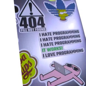 K&ouml;nnte beinhalten: Ein Laptop mit Aufklebern. Aufkleber sind ein 404-Fehler, ein Cartoon von Marge Simpson und Text mit der Aufschrift "I HATE PROGRAMMING" und "IT WORKS!" Eine rosa Cartoonfigur benutzt einen Laptop.