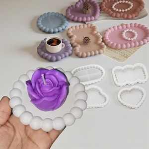 Puede incluir: Una colección de bandejas y moldes decorativos en varias formas y colores. Las bandejas son en tonos de rosa, morado y azul, con un borde de cuentas. Una vela morada en forma de rosa se encuentra en una bandeja blanca con cuentas. También, una taza de café.