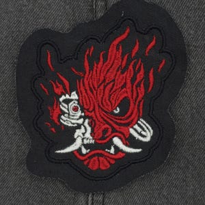 Op de afbeelding: Zwarte en rode geborduurde patch van een gestileerd demonen gezicht met vlammen. De demon heeft witte tanden en een rode tong.