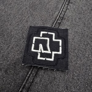 May include: Black denim fabric with a white embroidered patch of a stylized cross with a letter "M" inside.