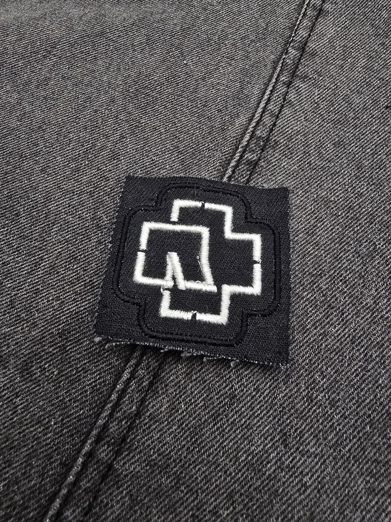May include: Black denim fabric with a white embroidered patch of a stylized cross with a letter "M" inside.