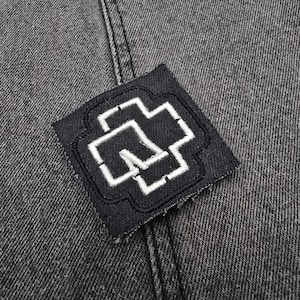May include: A black fabric patch with a white embroidered cross design. The patch is sewn onto a dark gray denim fabric.