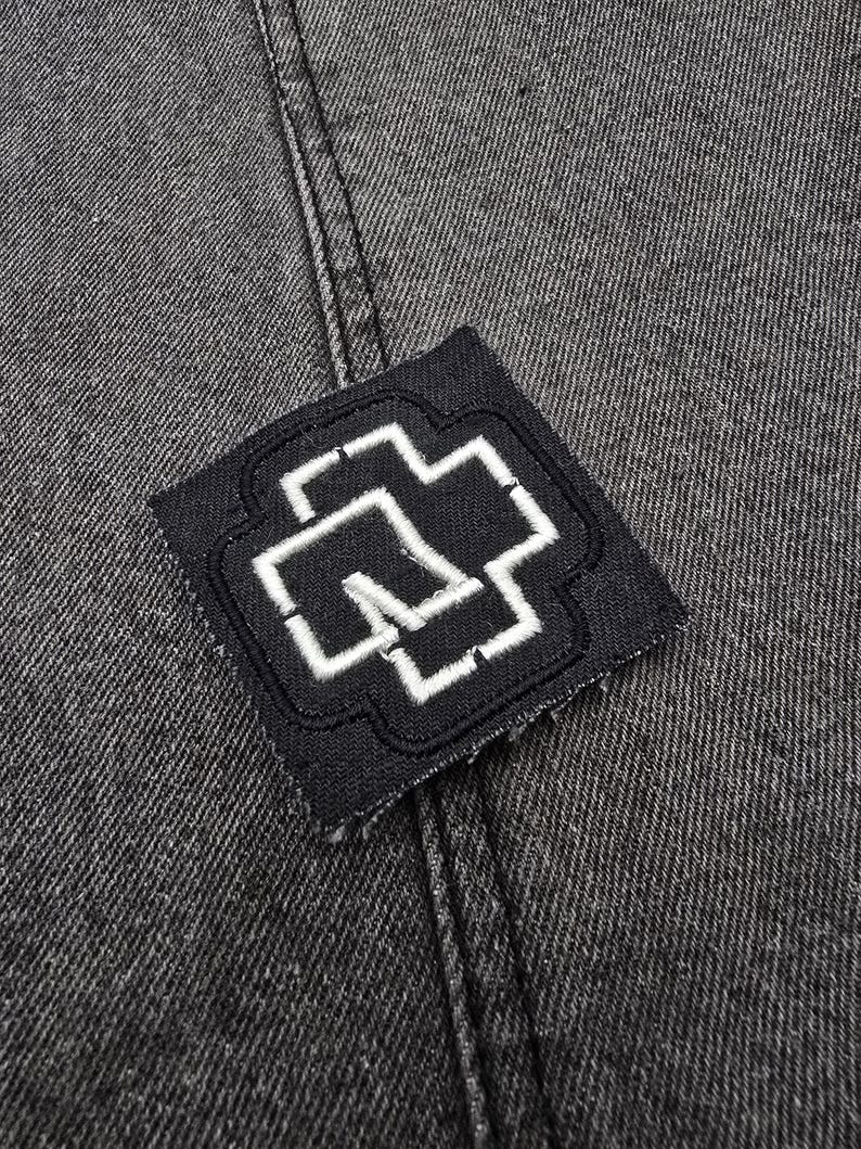 May include: A black fabric patch with a white embroidered cross design. The patch is sewn onto a dark gray denim fabric.