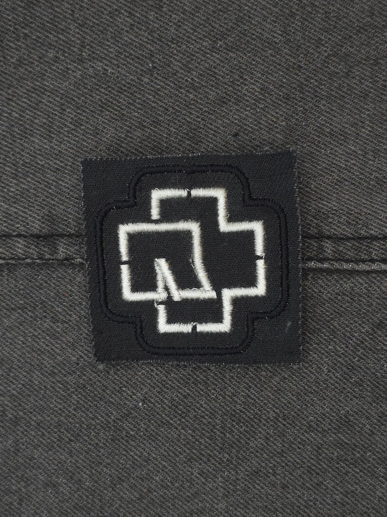 May include: Black and white embroidered patch with a stylized cross design. The cross is outlined in white and has a white shape inside.