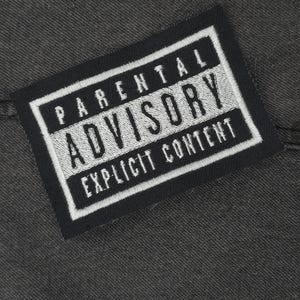 Op de afbeelding: Zwart-wit Parental Advisory-label met de tekst "Expliciete inhoud" op een donkergrijze stoffen achtergrond.