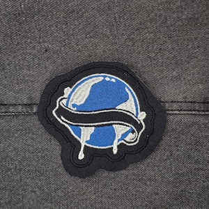 Op de afbeelding: Een geborduurde patch in zwart en blauw van een druppelende wereld met een zwarte banner eromheen.