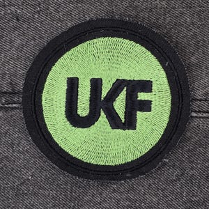 Op de afbeelding: Een geborduurde zwarte en groene patch met de letters "UKF" in een cirkel.
