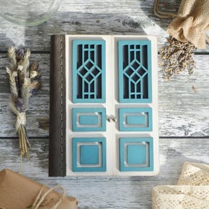 Puede incluir: Una caja de madera blanca con un diseño de puerta pintada de azul. La puerta tiene un patrón geométrico y está dividida en cuatro secciones. La caja está cerrada y tiene un ribete de cuero marrón.