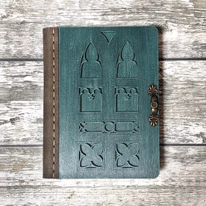 Puede incluir: Un diario de cuero verde azulado con un cierre de metal decorativo. La portada presenta un diseño tallado de una puerta arqueada con detalles intrincados.