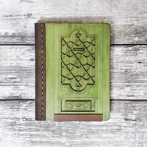 May include: A green leather-bound journal with a decorative, carved wooden cover featuring a geometric pattern. The journal has a brown leather spine and a brown leather flap.