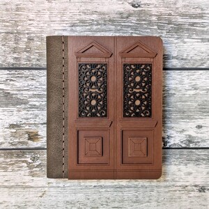 Puede incluir: Un diario encuadernado en cuero marrón con una cubierta de madera decorativa que presenta un diseño tallado de una puerta doble con detalles intrincados. La cubierta tiene un aspecto desgastado y un borde cosido.